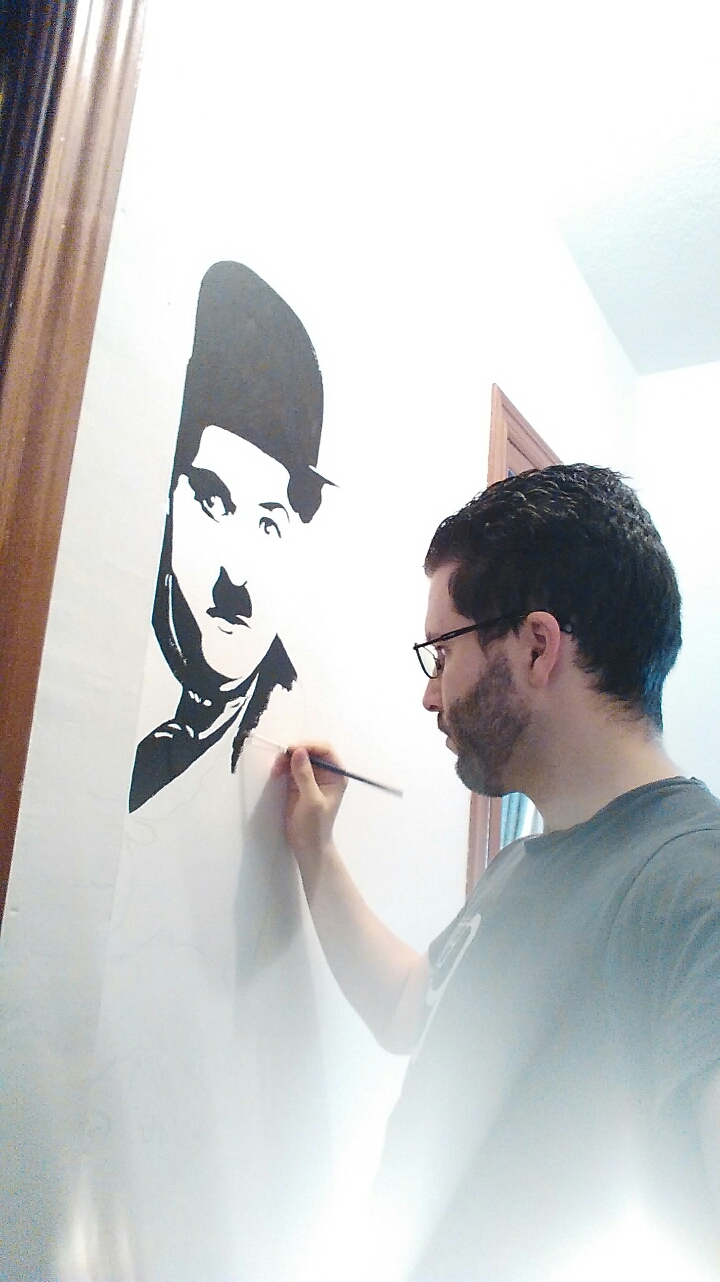 Fotografía del artista pintando un mural de Charles Chaplin en acrílico sobre pared blanca, proceso creativo en acción.